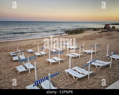 Blick auf den Sonnenuntergang am Strand mit weißen Sonnenliegen in Olhos de Agua, Algarve, Portugal. Stockfoto