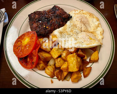 Frühstück mit Steak, Spiegeleiern, gebratenen Tomaten und Bratkartoffeln auf einem weißen Teller Stockfoto