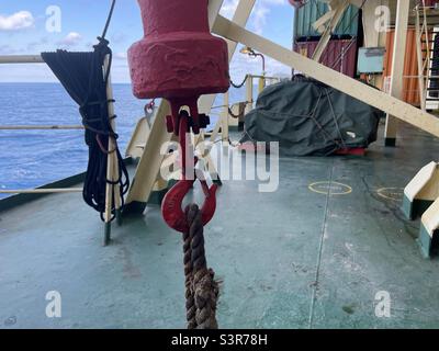 Nahsicht auf rotem Haken vom Bunker-Kran, der per Seil an das grüne Deck des Handelscontainers befestigt ist. Im Hintergrund ist ruhiges Meer, blauer Himmel und verstaute Container. Stockfoto