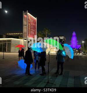 LOS ANGELES, CA, OCT 2021: Anonyme Menschen mit beleuchteten mehrfarbigen Regenschirmen im Music Center bei Nacht, mit dem Rathaus, das lila im Hintergrund leuchtet Stockfoto