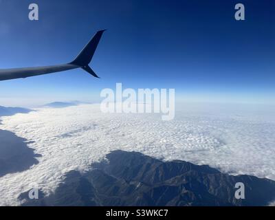 Blick vom Verkehrsflugzeug mit Flugzeugflügel, vom Flugzeugfenster aus gesehen, während man über Berge fliegt, die über niedrigen Wolken aufsteigen Stockfoto