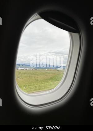 Berge, Wolken und ein entferntes Flugzeug, das durch das Passagierjetfenster gesehen wird, während es auf die Start- und Landebahn am Flughafen Missoula Montana rollt Stockfoto