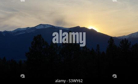 Dramatische Umrisse von Bergen in der Dämmerung, mit Sonnenuntergang hinter ihnen und dunklen, silhouettierten Kiefern kaum sichtbar im Vordergrund. Montana, USA Stockfoto