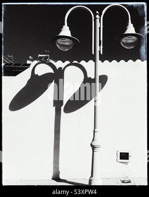 Helles Sonnenlicht wirft einen dunklen Schatten von Straßenlampen auf eine weiße Wand. Das Foto ist in Schwarz-Weiß gehalten. Stockfoto