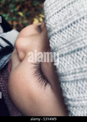 Schlafendes kleines Mädchen in einem grauen Wollhut, das in einer Schlinge auf einem Waldspaziergang getragen wird Stockfoto