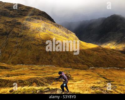 Weibliche Walkerin mit Lichtwellen, während die Wolken auf dem Pfad und den Hängen von Munro Buachaille Etive Beag, Glencoe, Schottland, zu brechen beginnen Stockfoto