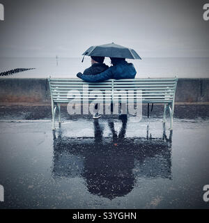 Das Paar sitzt im Regen auf einer Bank und blickt aufs Meer Stockfoto