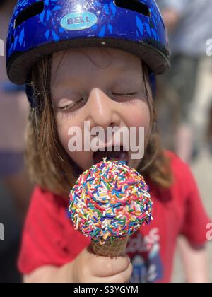 Fünf Jahre alter Junge trägt einen Fahrradhelm und genießt einen Eiskegel, der mit bunten Prise bedeckt ist. Stockfoto