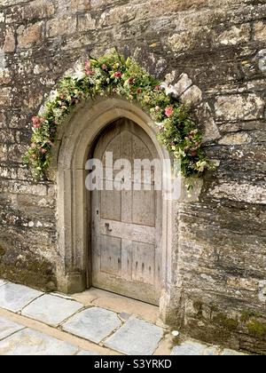 Wunderschöne hübsche hölzerne Kirchentür mit Steinbogen und wunderschöner Blumendekoration über der Tür für eine Hochzeit, St. Just in Roseland, Cornwall, England Stockfoto