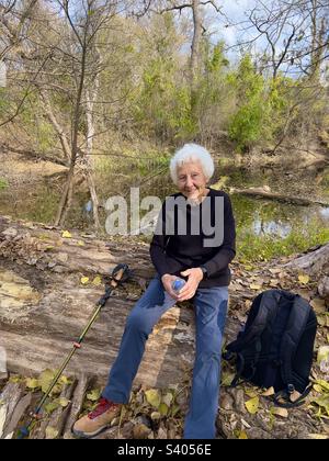 Wunderschöne Seniorin mit weißen Haaren, die nach der Wanderung im Herbst auf einem Baumstamm sitzt, mit all den Herbstblättern und wunderschönen Farben in Texas mit einem Teich im Hintergrund Stockfoto