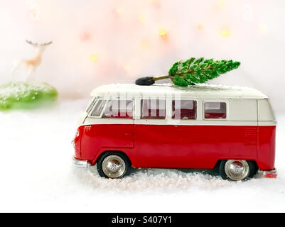 Der rote Camper fährt durch eine verschneite Szene mit einem Weihnachtsbaum auf dem Dach und beobachtet von einem Spielzeughirsch auf einem Hügel in der Ferne. Stockfoto