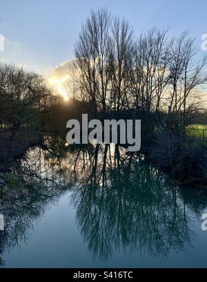 Winterliche Flussszene mit Bäumen, die sich im Wasser spiegeln, während die Sonne hinter Bäumen untergeht Stockfoto