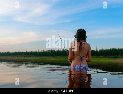 Ein Fischermädchen im Badeanzug steht bis zur Taille im Wasser des Flusses und fängt bei Sonnenuntergang mit einer Angel Fische Stockfoto