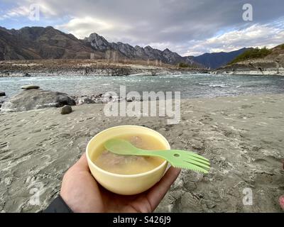 Eine Touristenhand hält eine Plastiksuppe mit Fleisch mit einer Gabel und einem Löffel vor dem Hintergrund des Flusses und der Berge in Altai. Stockfoto