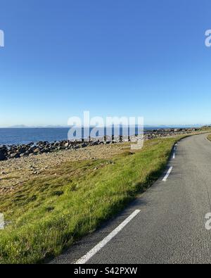 Straße am Meer auf den Lofoten-Inseln, Norwegen. An einem späten, sonnigen Sommerabend gedreht. Man kann auch Berge auf anderen Inseln weiter entfernt im Meer sehen. Stockfoto