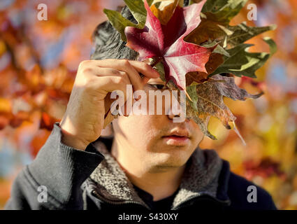 Porträt eines jungen Asiaten, der ein Herbstblatt vor seinem Gesicht hält. Stockfoto