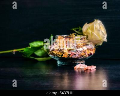 Nahaufnahme einer einzelnen gelben Rose und Potpourri in einem Glasbehälter mit getrockneten Blütenblättern auf schwarzem Hintergrund. Stockfoto