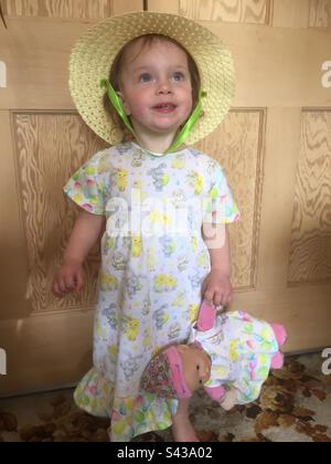 Kleinkind in einem hausgemachten Osterkleid aus Häschen und Eiern. Trägt eine gelbe Osterhut-Haube und hält eine Puppe in einem passenden Kleid Stockfoto