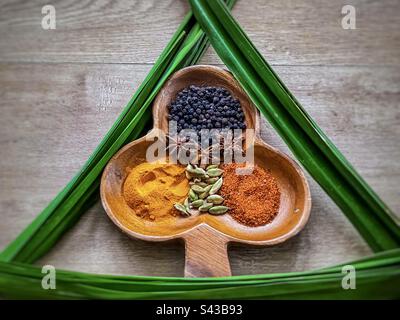 Flache Lage aus bunten Gewürzen in dekorativer Holzschüssel, eingerahmt von grünen Pandanusblättern, auch bekannt als Pandan- oder Schraubenpalmenblätter auf Holztisch. Stockfoto