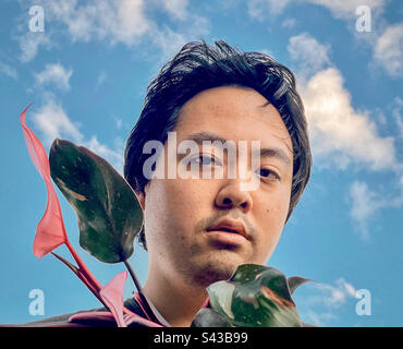 Kleinwinkelfoto eines jungen Asiaten mit rosa Prinzessin Philodendron gegen weiße Wolken am blauen Himmel. Stockfoto