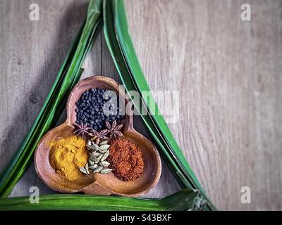 Direkt über der Aufnahme farbenfroher asiatischer Gewürze in einer dekorativen Holzschüssel, eingerahmt von grünen Pandanusblättern, auch bekannt als Pandan- oder Schraubenkiefernblätter auf Holz mit Kopierbereich. Stockfoto