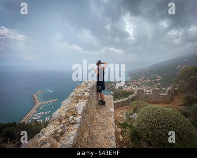 Ein Mann, der mit dem Telefon ein Foto von der unglaublichen Aussicht macht, steht auf einer alten Steinmauer, Rocca di Cefalù, Sizilien. Stockfoto