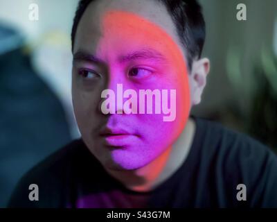 Nahaufnahme eines jungen Asiaten mit farbigem LED-Licht, das die Hälfte seines Gesichts vor unscharfem Hintergrund beleuchtet. Stockfoto