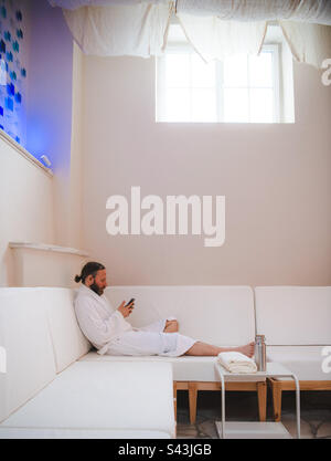 Ein Mann, der sich im Bademantel auf einer weißen Couch mit seinem Smartphone entspannt Stockfoto