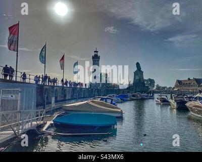 Silhouetten von Wanderern im Hafen von Lindau am Bodensee mit vielen geparkten Booten, Flaggen, Leuchtturm und Löwenstatue. Deutschland. Stockfoto