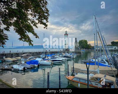 Blick auf den Sonnenuntergang am Hafen von Lindau mit Booten, Leuchtturm und Bergen am Bodensee in Deutschland. Stockfoto