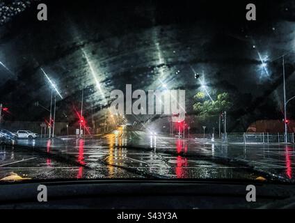 Blick auf die Straßenkreuzung, Ampeln, Autos und Ampeln durch die Windschutzscheibe des Autos in einer regnerischen Nacht. Stockfoto
