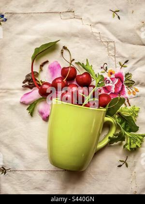 Blick aus der Vogelperspektive auf Kirschen, Salatblätter und Blüten, die aus dem grünen Becher auf einer alten bestickten Tischdecke herauslaufen. Saure Farbpalette. Textur. Textil. Kontrast, Nebeneinander. Stockfoto