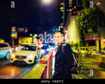 Ein junger asiatischer Mann mit Brille, der nachts auf dem Gehweg gegen Neonlichter, Gebäude und eine befahrene Straße in der Stadt steht. Selektiver Fokus. Farbenfroh. Stockfoto
