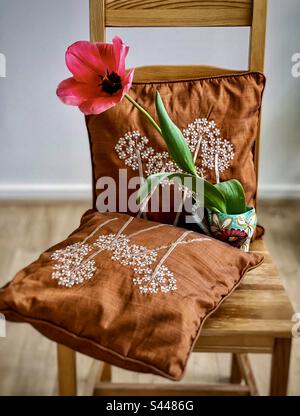 Eine pinkfarbene Tulpenblume in Vase mit gestickten Kissen auf einem Holzstuhl im Wohnzimmer. Böhmischer rustikaler Stil. Frühjahrsthema. Stockfoto