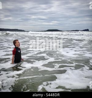 Ein neunjähriger Junge springt in das kalte Wasser und trotzt den großen Wellen in einem Neoprenanzug in der Irischen See am Rhossili Llangennith Beach in West Wales Stockfoto