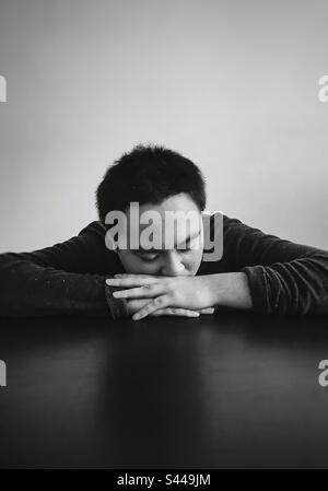 Monochromes Porträt eines jungen Asiaten mit geschlossenen Augen und Kopf auf gefalteten Armen auf dem Tisch. Speicherplatz kopieren. Emotionen. Traurigkeit. Müdigkeit. Stockfoto