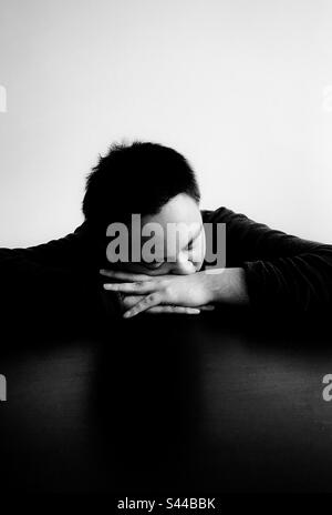 Nahaufnahme eines jungen Asiaten mit geschlossenen Augen und Kopf auf gefalteten Händen auf dem Tisch. Schwarzweiß. Speicherplatz kopieren. Dunkelheit. Emotionen. Traurigkeit. Stockfoto