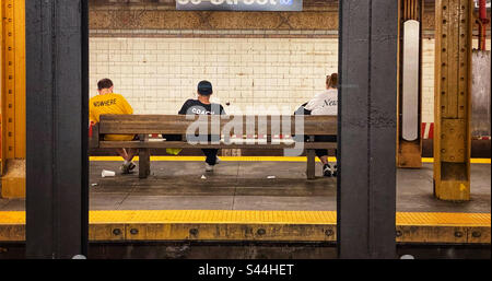 Drei Leute mit Wörtern auf ihren T-Shirts, die auf einer Bank sitzen und in einer U-Bahn-Station auf einen Zug warten Stockfoto