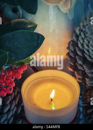 Blick aus einem hohen Winkel auf die beleuchtete Kerze und die Reflexion in Keramikfiguren, umgeben von Cotoneaster-Beeren und Pinienzapfen. Herbst-/Weihnachtsthemen. Vintage-Style. Spiritualität. Feier. Dekoration. Stockfoto