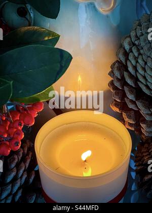 Blick aus einem hohen Winkel auf die beleuchtete Kerze und die Reflexion in Keramikfiguren, umgeben von Cotoneaster-Beeren und Pinienzapfen. Herbst-/Weihnachtsthemen. Vintage-Style. Spiritualität. Feier. Dekoration. Stockfoto