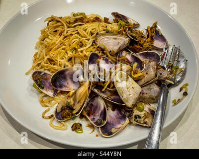 Nahaufnahme scharfer, unter Rühren gebratener Nudeln mit Pipis/Muscheln auf weißem Teller mit Besteck auf dem Tisch. Fusionsküche. Stockfoto