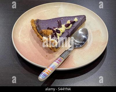 Blick aus einem hohen Winkel auf eine Scheibe Ube/Purple Yam Pie mit Vanilleschuhe und lila Yamswurzeln auf dem Teller mit einem Löffel auf dem Tisch. Traditionelles philippinisches Dessert. Stockfoto