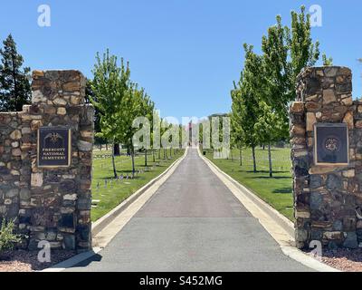 Prescott National Cemetery, Blick auf das historische Eingangstor, Center Drive führt zu American Flag, Veteranengräbern mit amerikanischen Flaggen, leuchtend blauem Himmel, eingerahmt von grünen Bäumen, Arizona, USA Stockfoto