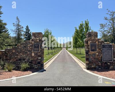 Prescott National Cemetery, Blick auf das historische Eingangstor, Center Drive führt zu American Flag, Veteranengräbern mit amerikanischen Flaggen, leuchtend blauem Himmel, eingerahmt von grünen Bäumen, Arizona, USA Stockfoto