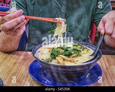 Mittelteil eines Mannes, der mit Stäbchen Vermicelli-Nudeln aus der Schüssel dampfender heißer Vermicelli-Suppe auf dem Tisch hebt. Stockfoto