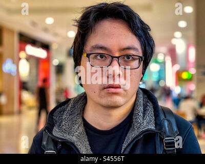 Nahaufnahme eines jungen Asiaten mit Brille gegen Lichter im Einkaufszentrum. Konzentrieren Sie sich auf den Vordergrund. Stockfoto