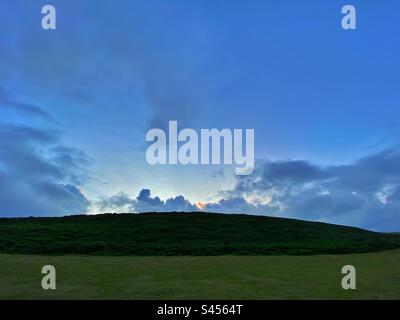 Ein Foto einer Landschaft - ein grünes Feld und ein Hügel mit einem versteckten Sonnenaufgang. Blauer Himmel, Kopierraum Stockfoto