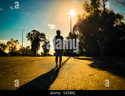 Rückansicht eines Mannes, der gegen Bäume, Lampenpfähle, untergehende Sonne und Himmel während des Sonnenuntergangs geht. Hintergrundbeleuchtet. Silhouette. Goldene Stunde. Stockfoto