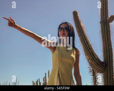 Junge, langhaarige, lächelnde Frau in Sonnenbrille und gelbem Kleid, die mit erhobenem Arm neben einem großen Kaktus vor klarem, blauem Himmel posiert. Mode. Glück. Spaß. Stockfoto