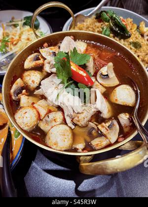 Aus einem hohen Blickwinkel bietet sich eine scharfe thailändische Tom Yum Suppe mit Pilzen, Hühnchen und Kräutern in einem heißen Topf, umgeben von verschiedenen thailändischen Gerichten auf dem Tisch. Stockfoto